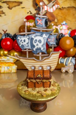Pirate-themed birthday cake shaped like a treasure chest on gold stand with skull ship decoration and colorful balloons backdrop.