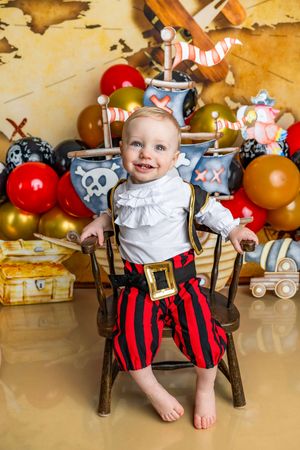 A smiling baby in a pirate costume sits on a wooden chair surrounded by colorful balloons and pirate-themed decorations.