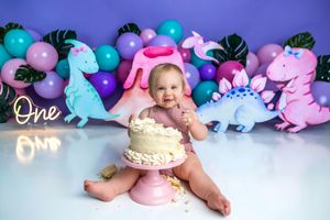 Baby smashing a white cake at a dinosaur-themed first birthday party with colorful balloons and dino cutouts.
