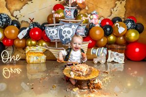 Baby smashing pirate-themed first birthday cake surrounded by gold, red, and black balloons with a treasure map backdrop.