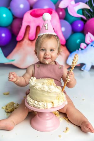 Baby girl in pink party hat smashes first birthday cake surrounded by colorful dinosaur balloon decorations.