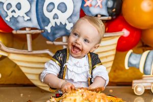 Baby in pirate costume smashing first birthday cake with chocolate frosting on face, pirate ship backdrop with balloons.