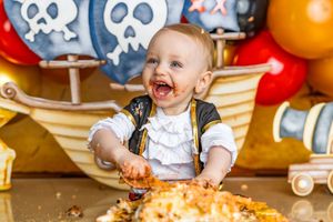 Happy baby in pirate costume smashing first birthday cake at a pirate-themed party with colorful balloons.