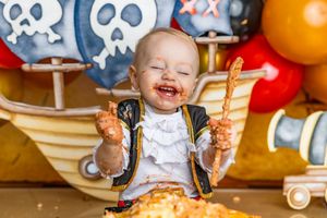 Baby smiling with cake-covered hands at a pirate-themed first birthday party with colorful balloons.