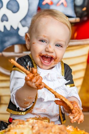 Messy baby in pirate costume laughing while smashing first birthday cake, covered in chocolate frosting.