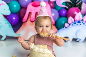 Baby girl in pink party hat eating first birthday cake surrounded by colorful dinosaur decorations and balloons.