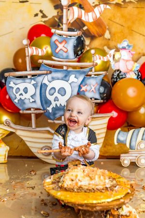 Laughing baby in pirate costume smashing first birthday cake at pirate-themed party with ship backdrop and colorful balloons.