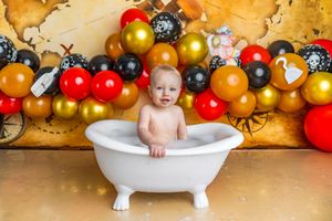 Baby sitting in a white mini bathtub surrounded by red, gold, and black balloons for a first birthday cake smash.