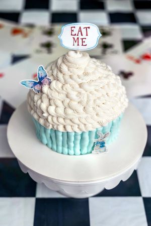 Alice in Wonderland giant cupcake with white frosting, teal wrapper, butterfly decoration, and an Eat Me topper on a cake stand.