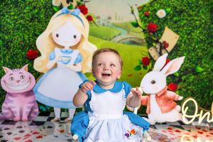 Baby girl in Alice in Wonderland dress smiling at first birthday cake smash photoshoot with themed backdrop.