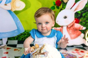 Baby in Alice in Wonderland costume smashing first birthday cake with White Rabbit backdrop.