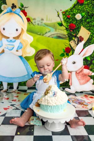 Baby smashing first birthday cake at Alice in Wonderland themed party with White Rabbit and Alice decorations.