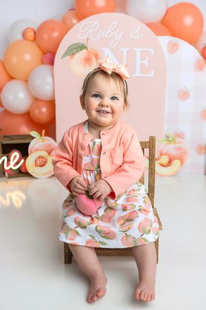 Baby girl in peach outfit sits on wooden chair at her first birthday party with balloon arch backdrop.