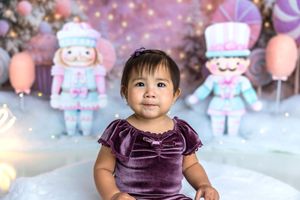 Baby girl in purple velvet dress sits before nutcracker decorations and pastel balloons for first birthday photo.