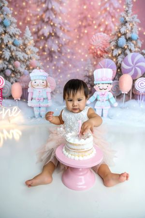 Baby girl in tutu smashes first birthday cake on pink stand with nutcracker and candy winter backdrop.