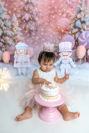 Baby girl in tutu smashes first birthday cake surrounded by pastel nutcracker decorations and pink trees.