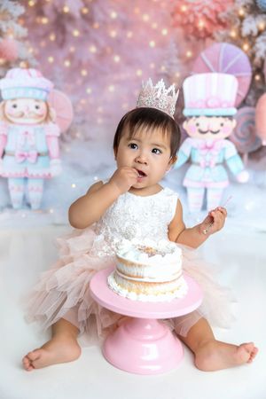 Baby girl in white dress and crown sits beside a pink cake stand for her first birthday cake smash photo.