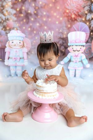 Baby girl wearing crown enjoys first birthday cake smash with nutcracker decorations in pink winter setting.