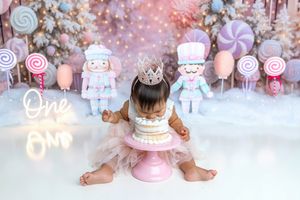 Baby girl in tutu sits before a candy-themed first birthday cake smash backdrop with nutcrackers and lollipops.