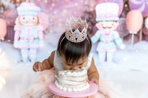 Toddler in pink tutu and silver crown leans into a white birthday cake during a cake smash session.