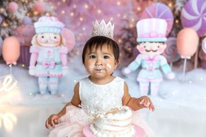 Adorable baby girl in white dress and crown sits with cake at a pastel nutcracker first birthday smash.