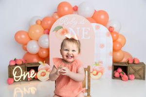Baby girl smiling at her first birthday party with peach balloon arch and ONE sign backdrop.