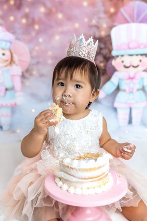 Adorable baby girl wearing a crown smashes her first birthday cake, face covered in frosting, pink tutu dress.
