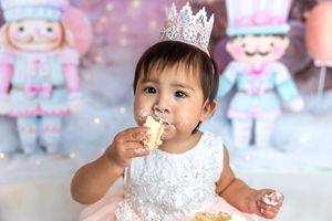 Baby girl in white dress and crown eats cake at a pink nutcracker-themed first birthday cake smash.