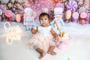 Baby girl in pink tutu and crown sits among candy decorations for a first birthday cake smash photo.