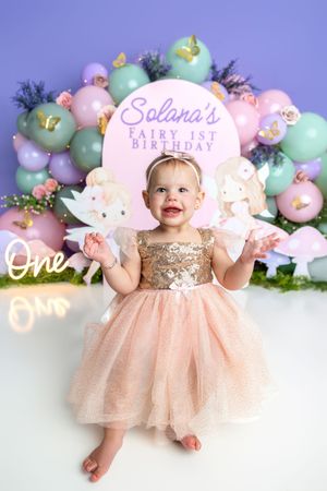 A baby girl in a gold sequin and pink tutu dress celebrates her fairy-themed 1st birthday with colorful balloon backdrop.