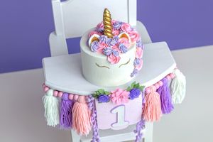 Unicorn smash cake on a white high chair with a garland banner showing number 1, decorated with pink and purple tassels.