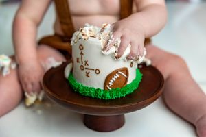 Baby smashing a football-themed first birthday cake on a wooden cake stand during a cake smash photoshoot.