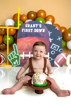 Baby Grant celebrates his first birthday with a football-themed cake smash, surrounded by brown, green, and white balloons.