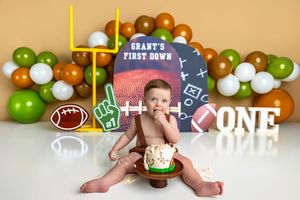 Baby boy enjoys his first birthday cake smash with a football theme, surrounded by colorful balloons and a Grant's First Down backdrop.