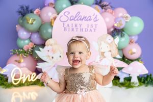 Baby girl in gold sequin dress celebrates fairy 1st birthday with pastel balloon arch and unicorn decorations.