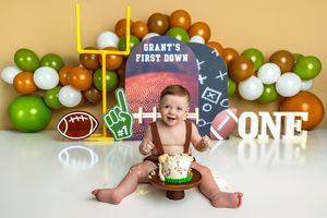 Baby boy celebrates first birthday with football-themed cake smash, surrounded by green, white, and orange balloons.