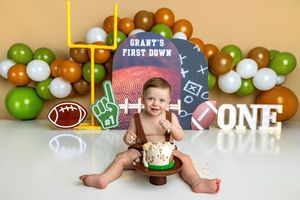 Baby boy celebrates first birthday with football-themed cake smash, surrounded by green, white, and brown balloons.