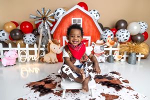 Smiling baby boy in cowboy bandana sits on cow print rug with farm barn backdrop and balloons for first birthday.