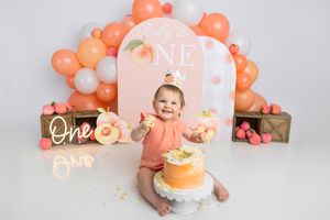 Baby girl smiles at her peach-themed first birthday cake smash with orange balloons and decorations.