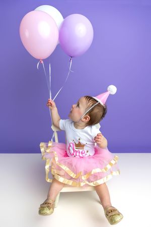 Baby girl in pink tutu and party hat holds colorful balloons on her first birthday celebration.