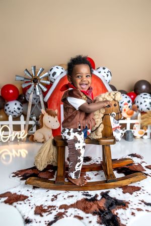 A smiling toddler in cowboy outfit sits on wooden rocking horse, surrounded by farm-themed balloons and decorations.