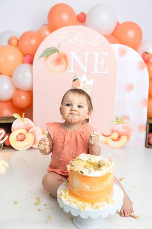 Baby girl Ruby celebrates her first birthday smashing a peach-themed cake at a peach balloon backdrop party.