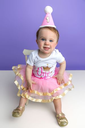 Baby girl in pink tutu and birthday hat celebrating her first birthday against a purple background.