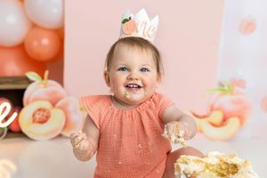Smiling baby girl with peach crown celebrates first birthday cake smash with frosting on her hands.