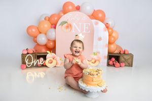 Baby girl in peach outfit laughs joyfully at her first birthday cake smash with peach balloon arch backdrop.