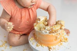 Baby in peach romper smashing first birthday cake, covered in frosting and crumbs during cake smash photoshoot.