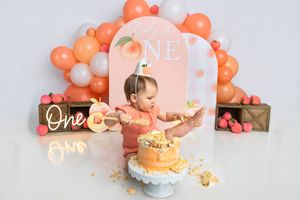 Baby girl celebrates first birthday smashing a peach cake surrounded by orange balloon decorations and ONE signs.