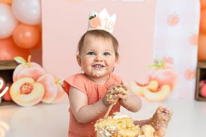 Baby girl in peach dress smashes first birthday cake, wearing crown, surrounded by peach balloons and decorations.