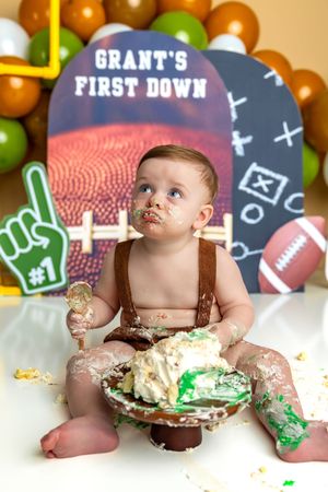 Baby Grant enjoys his first birthday cake smash with a football theme, surrounded by balloons and a 'First Down' sign.