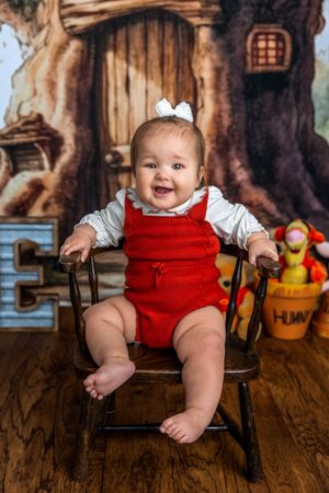 Smiling baby in red overalls sits on wooden chair with Winnie the Pooh backdrop and stuffed animals.